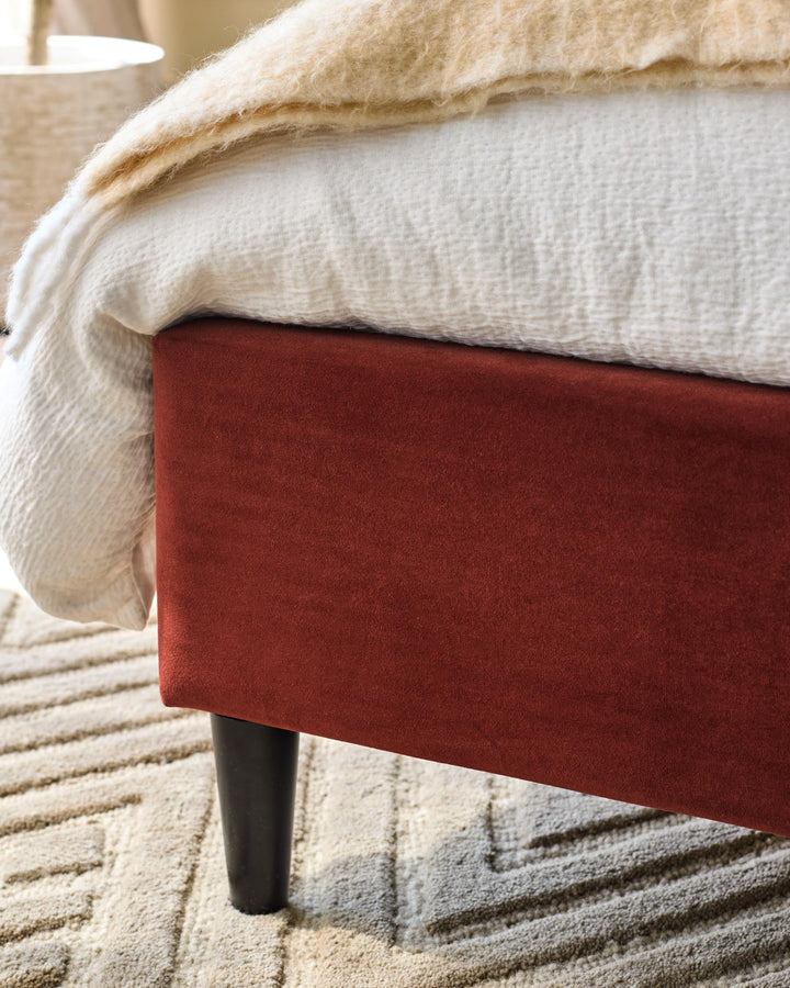 Close up of a bed with a dark red base, wooden legs and a cream textured throw and white bed sheet.