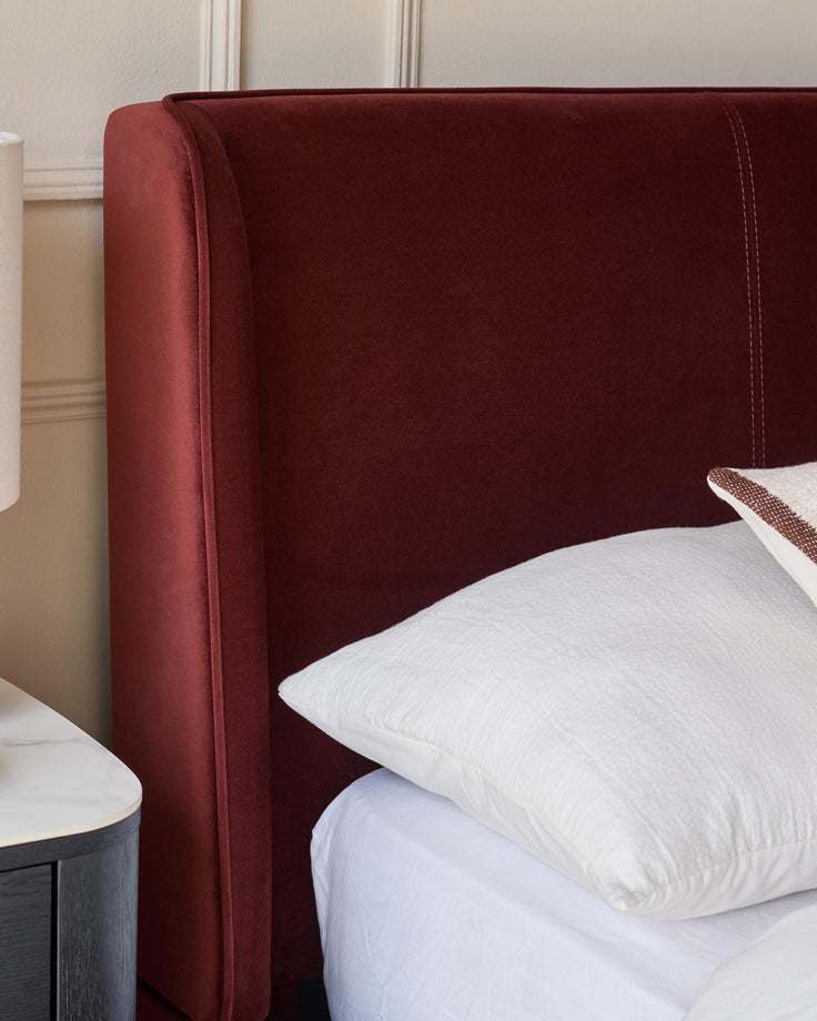 Close-up of a bedhead in a deep red velvet fabric, a white pillow on a white sheet, and a bedside table.