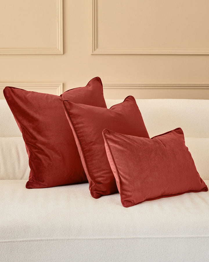 Luca cushion in medium toffee red, displayed on a cream bed against a beige paneled wall.