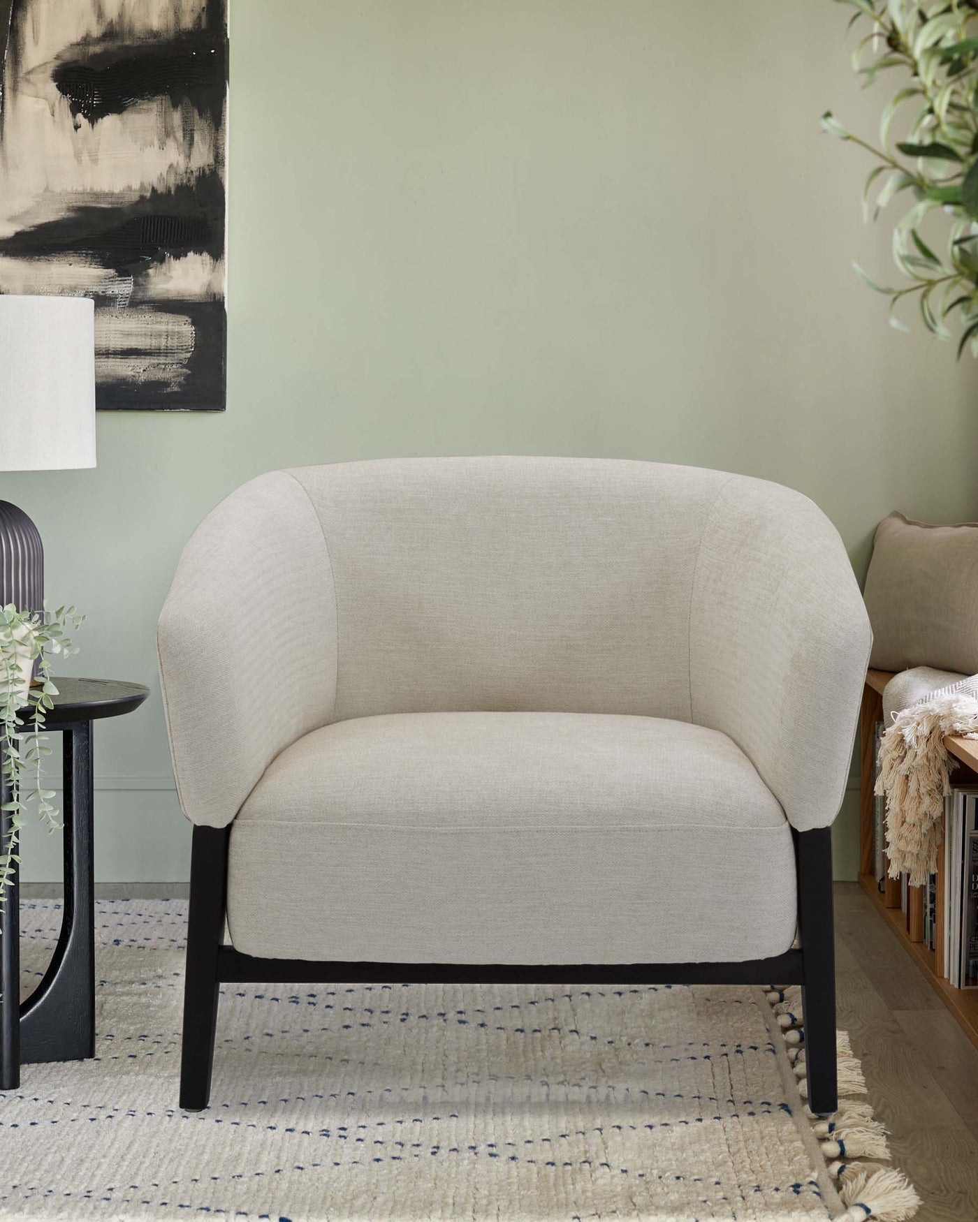 Modern light beige two-seater sofa with curved backrest and dark wooden legs, paired with a black round side table. The setting includes a textured beige rug with blue accents and a soft cream-colored tasselled throw on a bookshelf in the background.