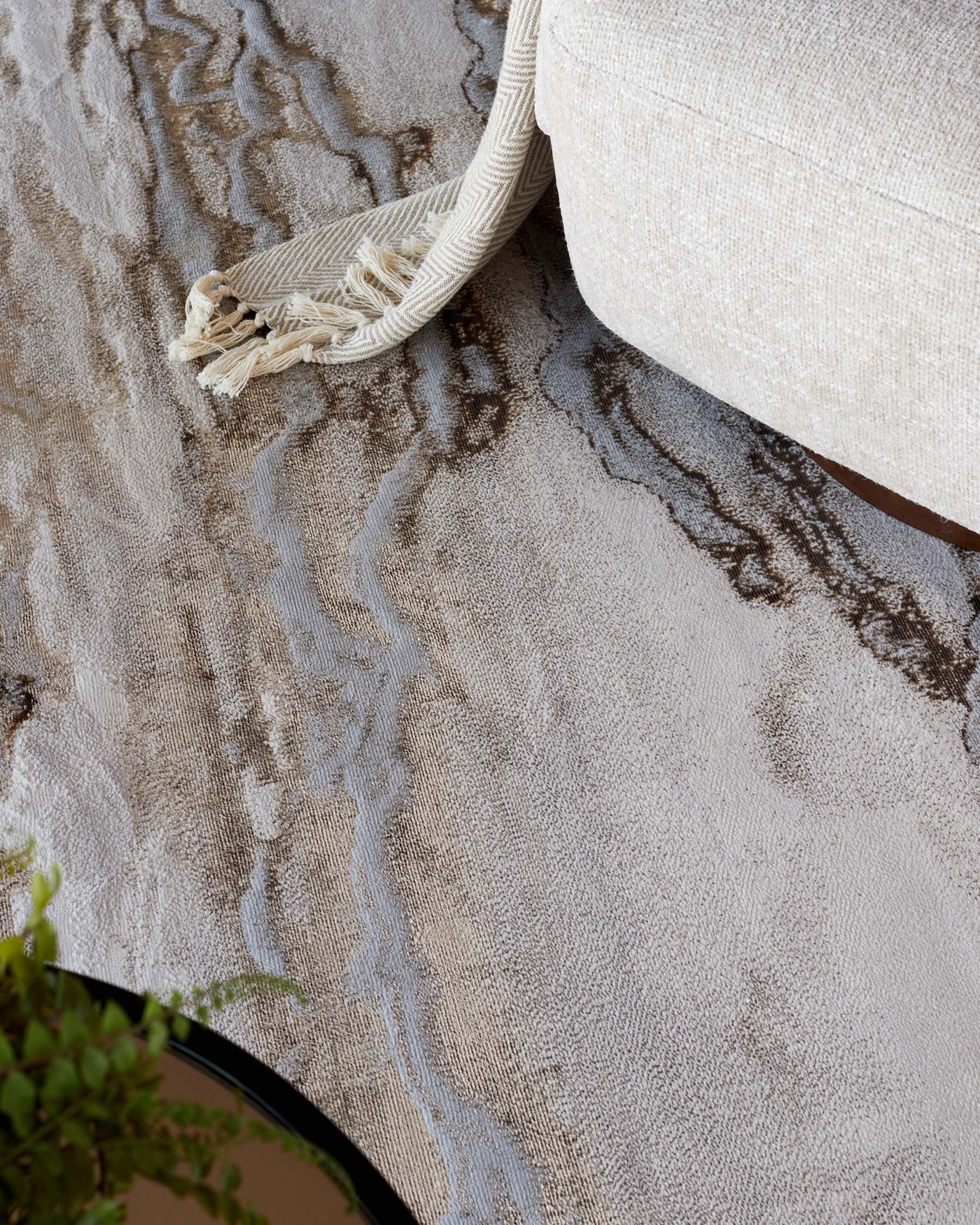 A close-up of a cream-colored fabric sofa with a textured throw blanket hanging over its side. Below is an abstract-patterned rug in shades of beige, cream, and dark brown with a hint of blue accent.