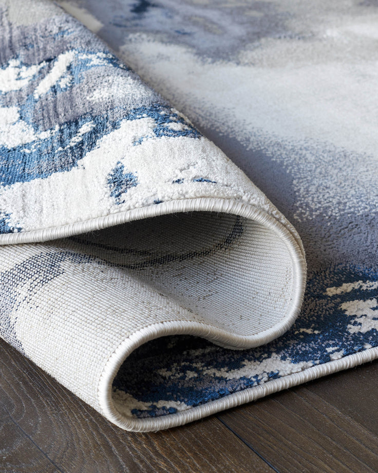 A modern rug with a blue, grey, and white abstract pattern is partially rolled up on a dark wooden floor.