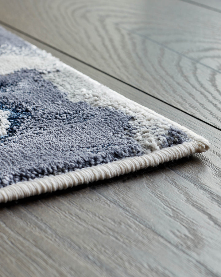 A corner of a patterned blue and grey rug on a wooden floor is displayed, showing the rug's texture and soft colours.