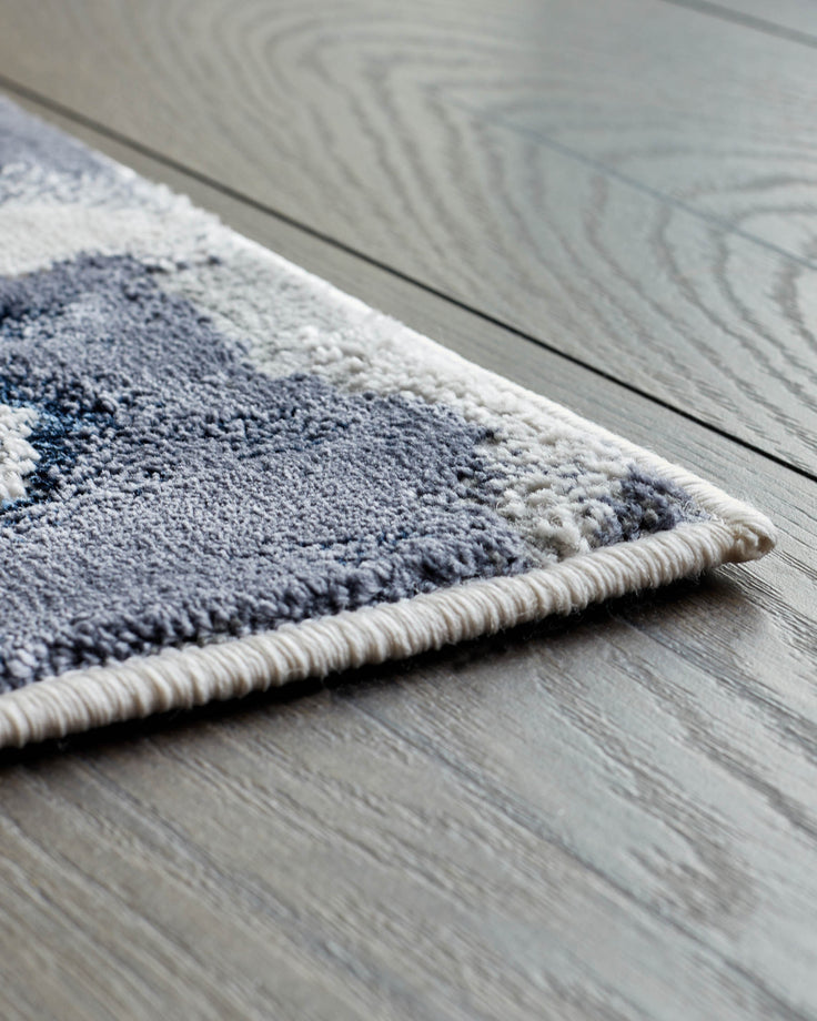 A corner of a patterned blue and grey rug on a wooden floor is displayed, showing the rug's texture and soft colours.