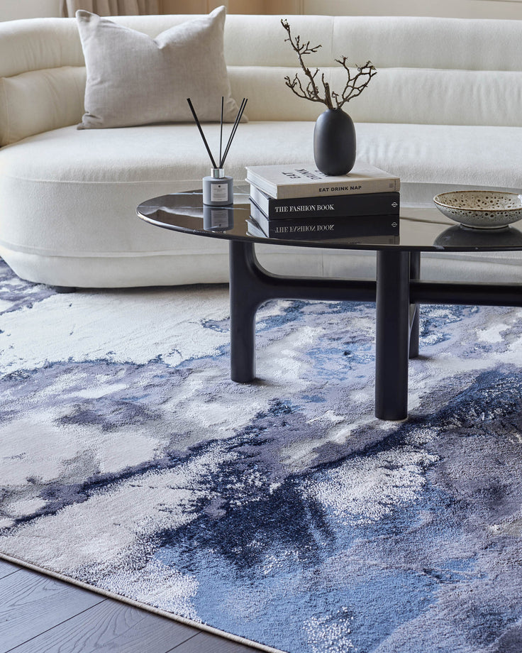 A modern living room featuring a curved sofa, glass-topped coffee table, stylish books, and an abstract patterned rug.