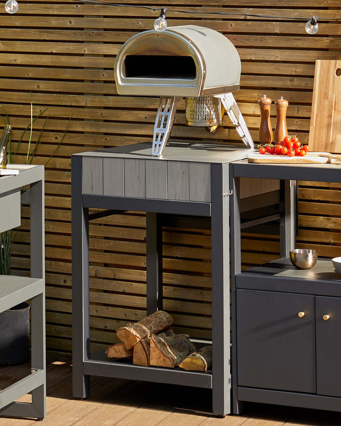 Outdoor kitchen furniture featuring a charcoal grey wood-fired pizza oven on a matching kitchen prep table with storage shelf and a side cabinet with two doors for storage. The scene is set against a wooden slatted background, suggesting a patio or garden environment ideal for outdoor cooking and entertainment.