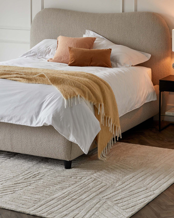 Modern upholstered bed with curved headboard in a textured beige fabric, featuring white bedding with a mix of beige and burnt orange pillows and a caramel-coloured fringe throw blanket. Beside the bed is a minimalist black nightstand with an open shelf. Under the bed lies a textured off-white area rug with a linear pattern.