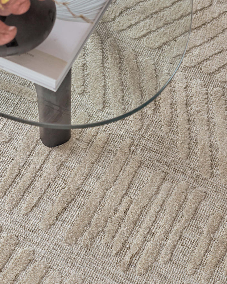Modern, minimalist glass coffee table with a single cylindrical dark leg and a round, clear tabletop. It rests on a textured beige rug with parallel line patterns.