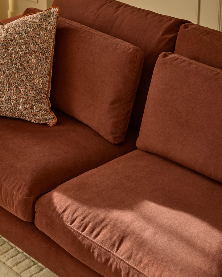 Quinn 2-seater sofa in toffee-red fabric with decorative pillows, shown from angled side view.