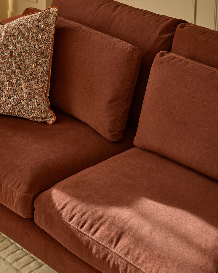 Quinn 2-seater sofa in toffee-red fabric with decorative pillows, shown from angled side view.