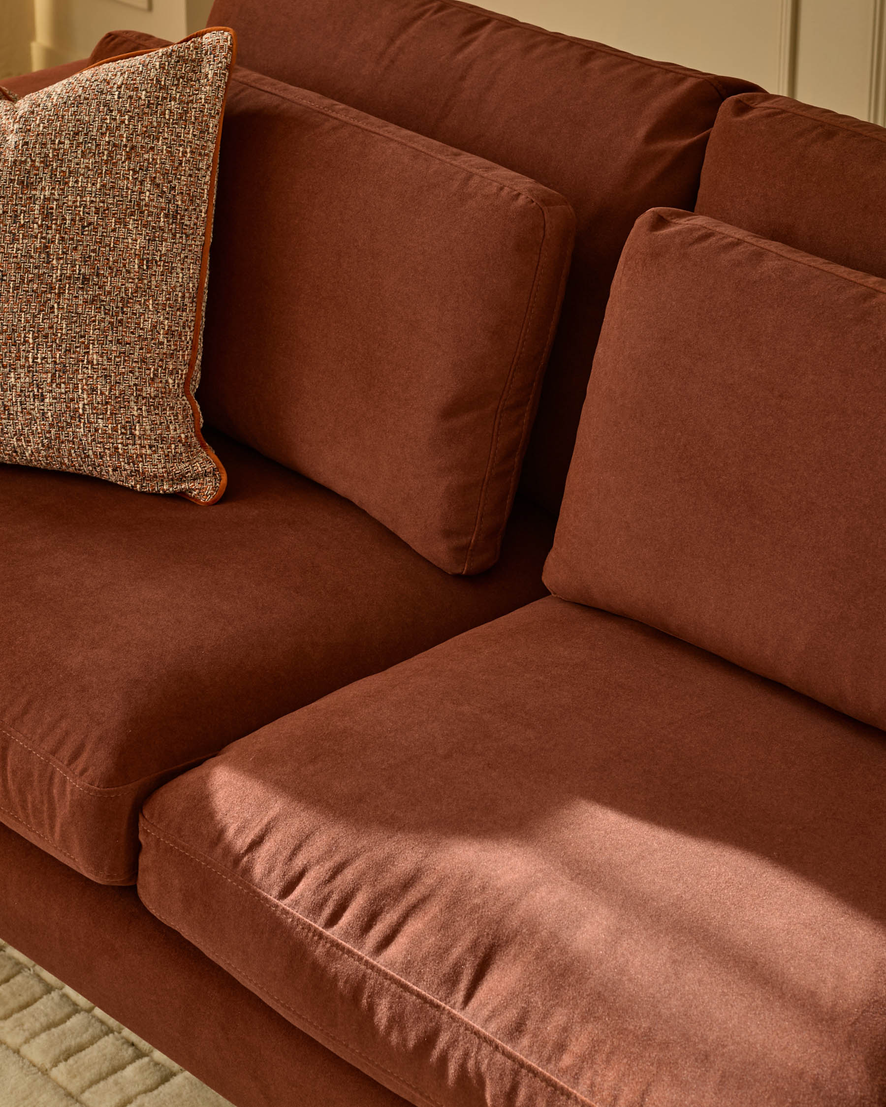 Quinn 3-seater sofa in toffee red fabric with decorative throw pillow, angled front view showing cushioned seats and back.