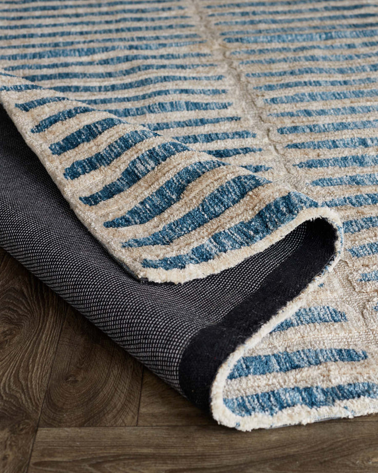 A striped rug with blue and beige bands, partially rolled up to reveal the backing, sits on a dark wood floor.