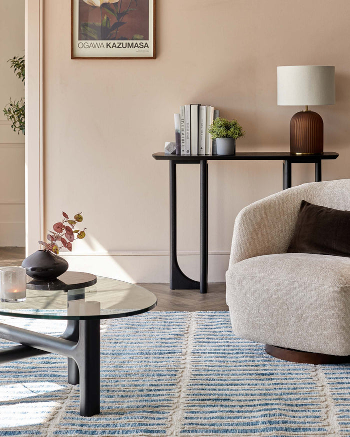 A modern living room showcasing a textured beige armchair with a dark brown pillow, a sleek round glass-top coffee table with a black base, and a slim black console table adorned with books, a metal lamp, and small potted plants, all resting atop a blue and beige striped area rug.