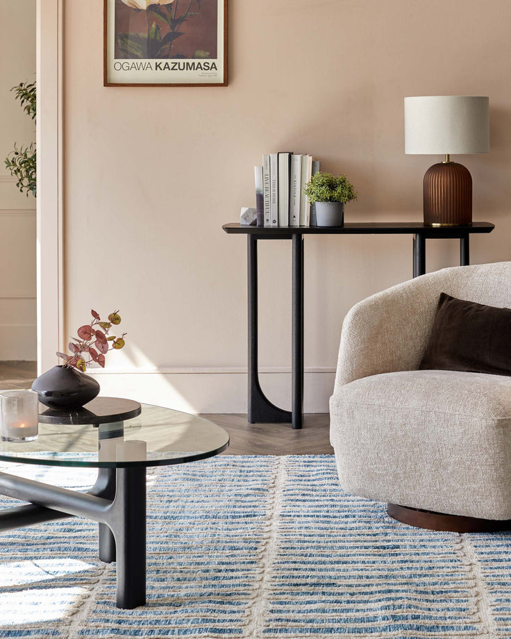 Living room furniture including a glass coffee table, side table with lamp, chair, and a patterned blue rug.