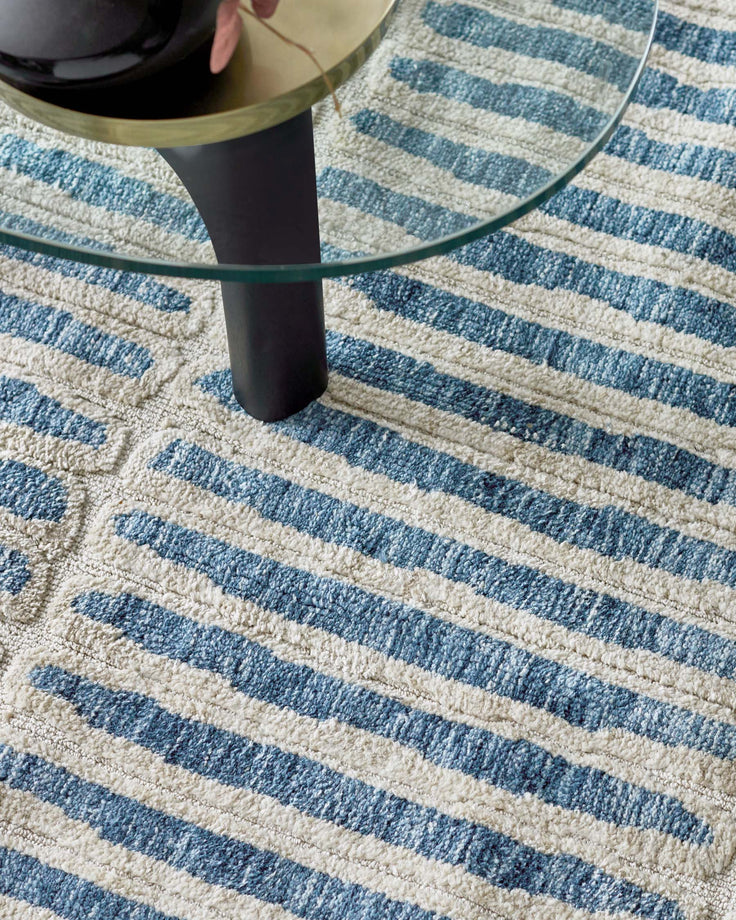 A round glass coffee table with a black, cylindrical base sits on a striped blue and cream rug.