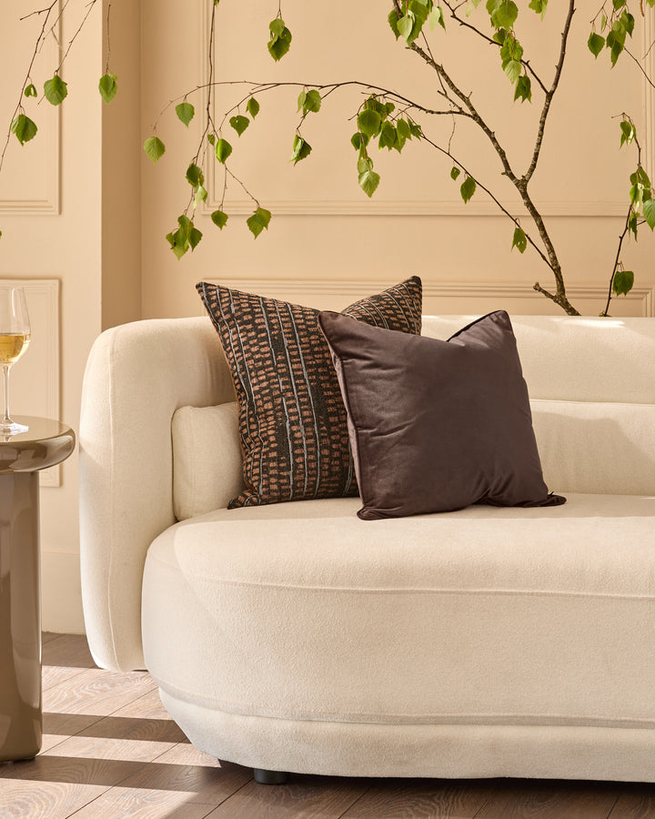 Luca medium chocolate cushion displayed on a cream armchair in a styled living room setting.