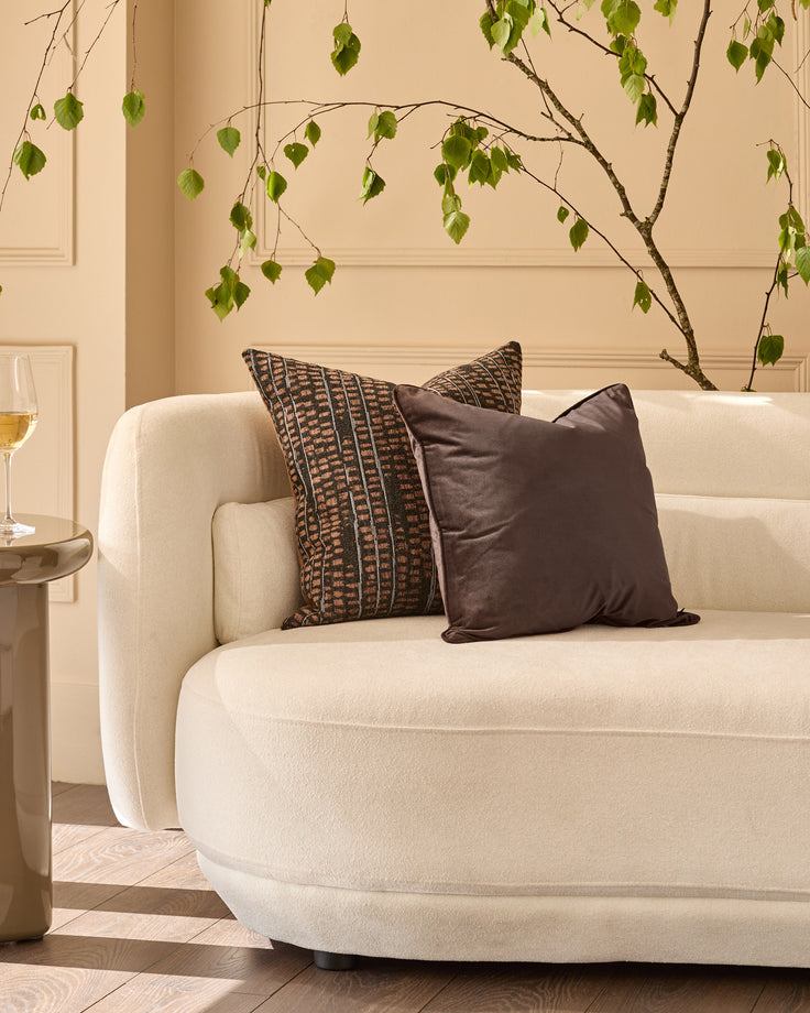 Luca medium chocolate cushion displayed on a cream armchair in a styled living room setting.