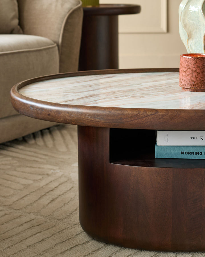 Rayna coffee table in dark wood with light marble top, cylindrical base, and open shelf displaying books.
