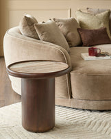 Dark wood round side table with marble top, positioned next to a curved beige upholstered sofa in a living room setting.