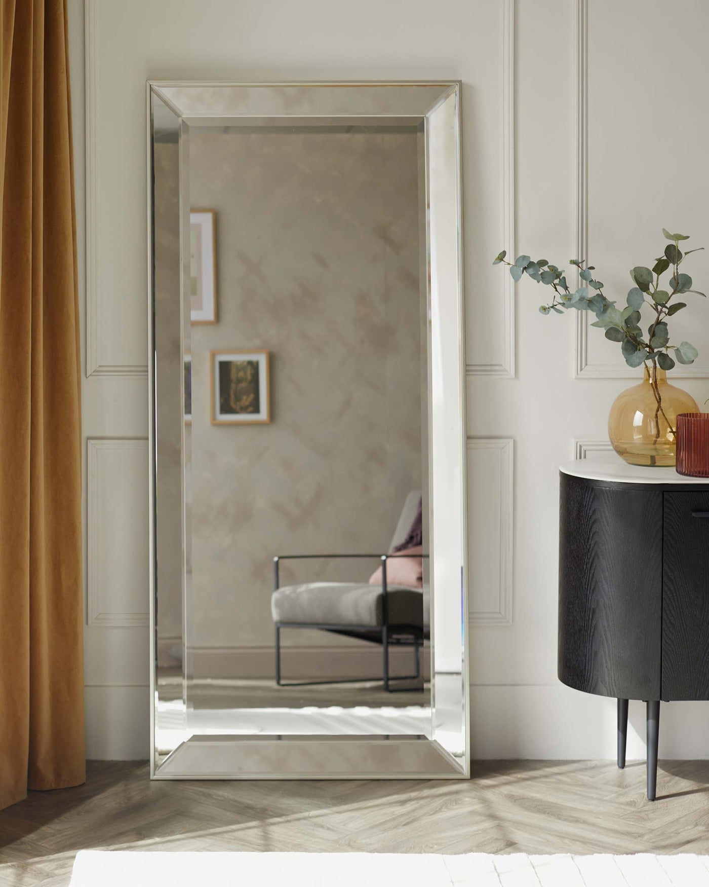 A large, floor-standing mirror with a sleek, bevelled frame, and a round, black contemporary sideboard with elegant, tapered legs. The sideboard is adorned with a glass vase holding eucalyptus branches and adjacent to it, a round, amber-coloured glass vase.