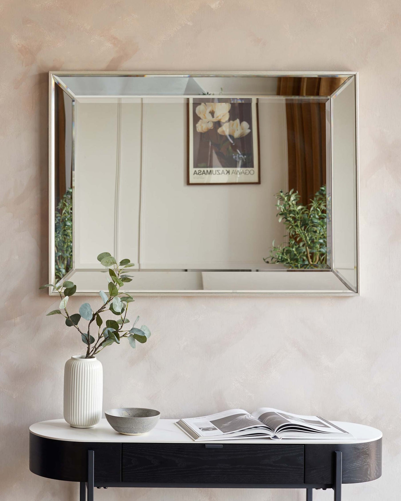 An elegant demilune console table with a black finish and sleek design, accompanied by a large rectangular wall mirror with a minimalist golden frame. Decor includes a textured white vase with greenery, a metallic bowl, and an open magazine.