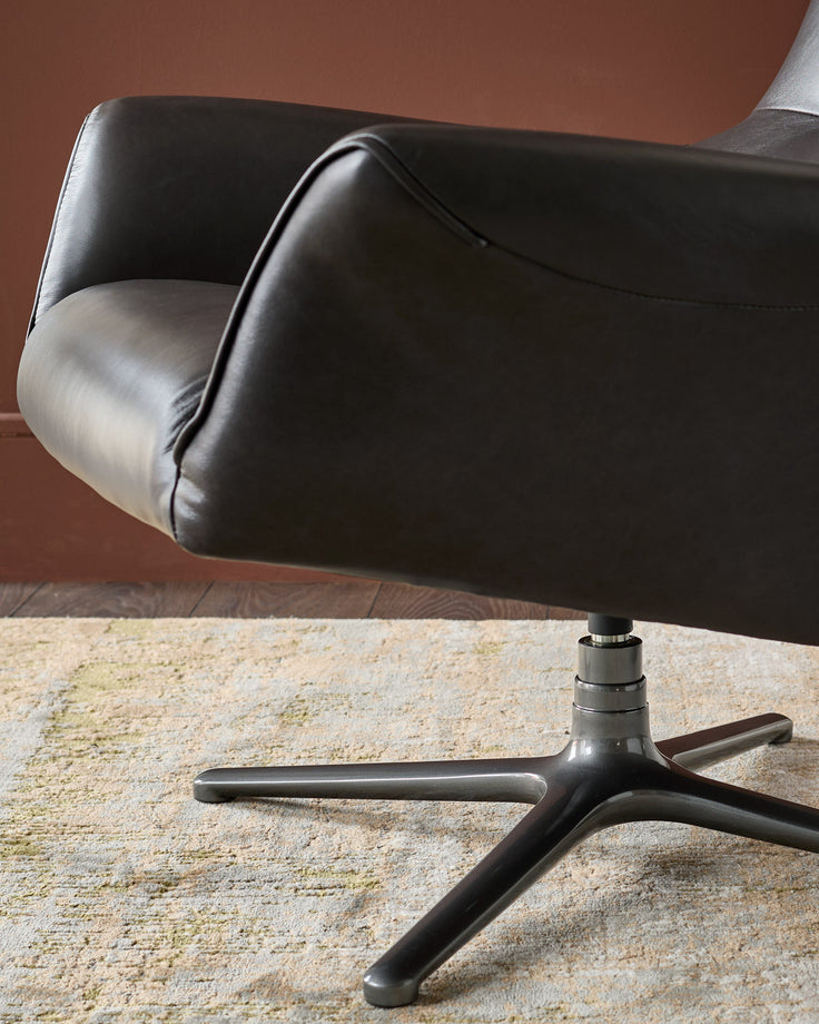 Black leather occasional chair with curved backrest and armrests, shown from side angle on three-leg metal base against brown wall.