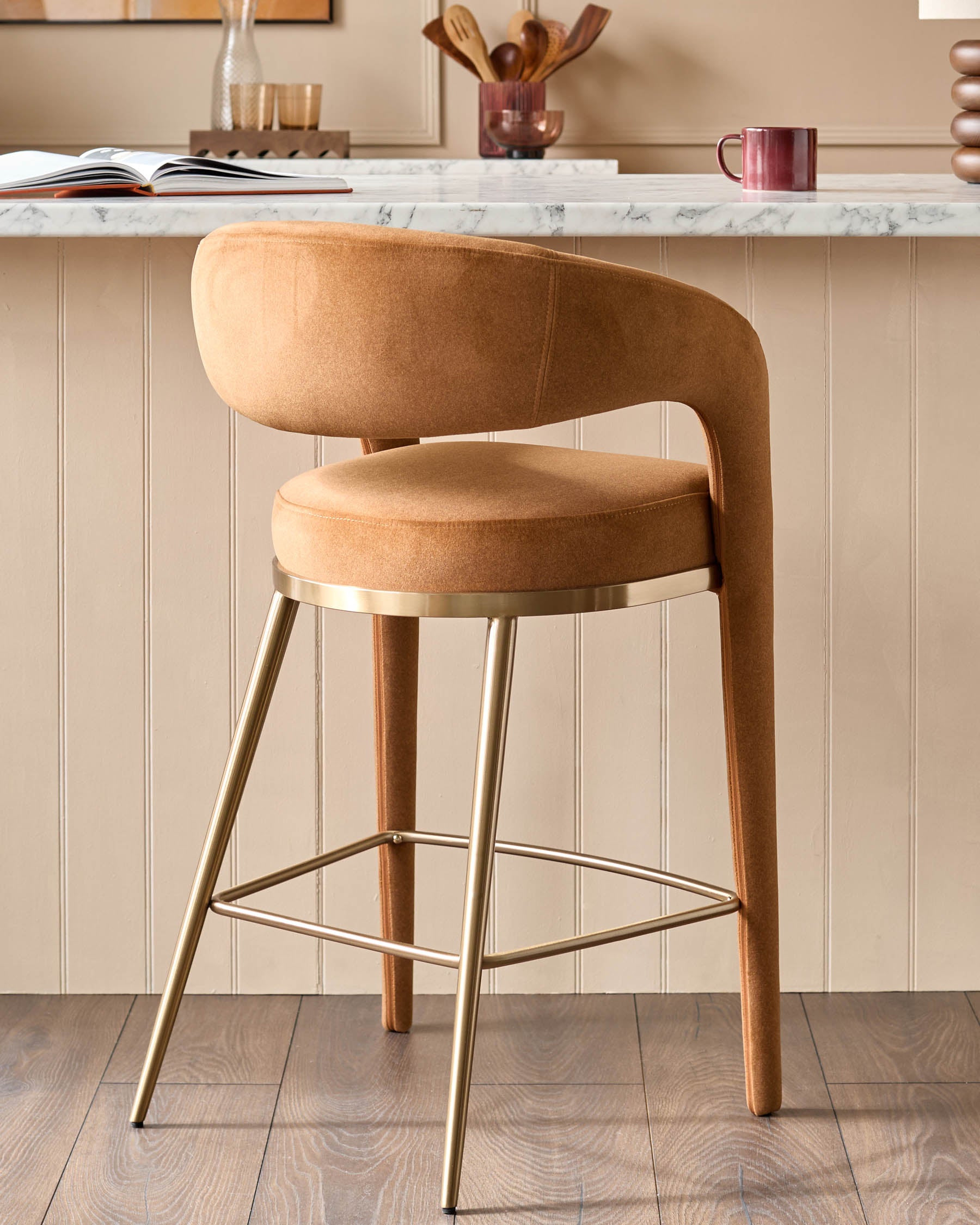 A modern bar stool with a gold frame and tan upholstery, set against a neutral kitchen backdrop.