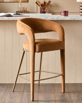 A stylish tan bar stool with a curved back and gold-coloured metal legs sits on a wooden floor in front of a neutral wall.