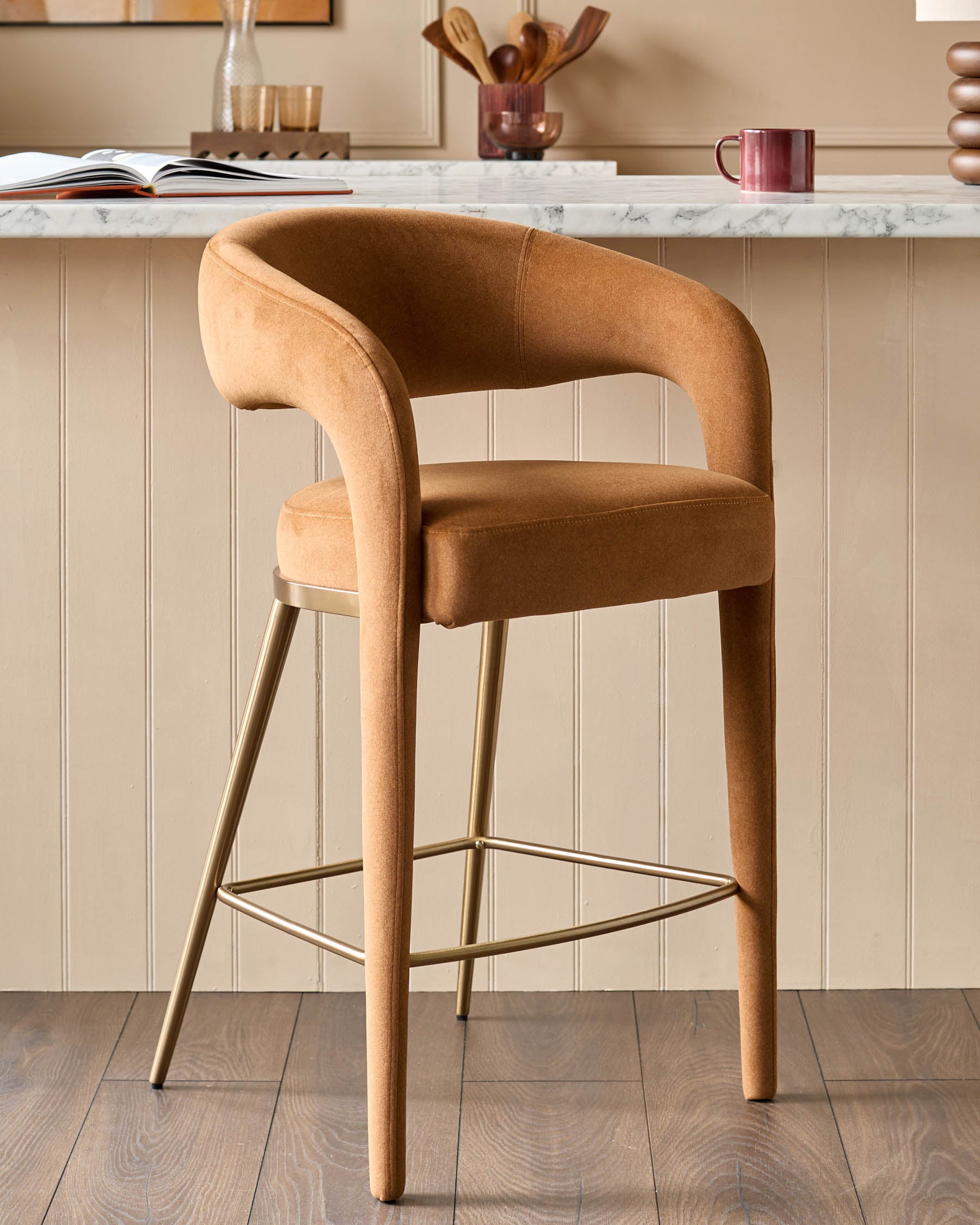 A stylish tan bar stool with a curved back and gold-coloured metal legs sits on a wooden floor in front of a neutral wall.