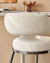 Close-up of a modern bar stool with a curved, cream-coloured seat and back, and a sleek, metallic base.