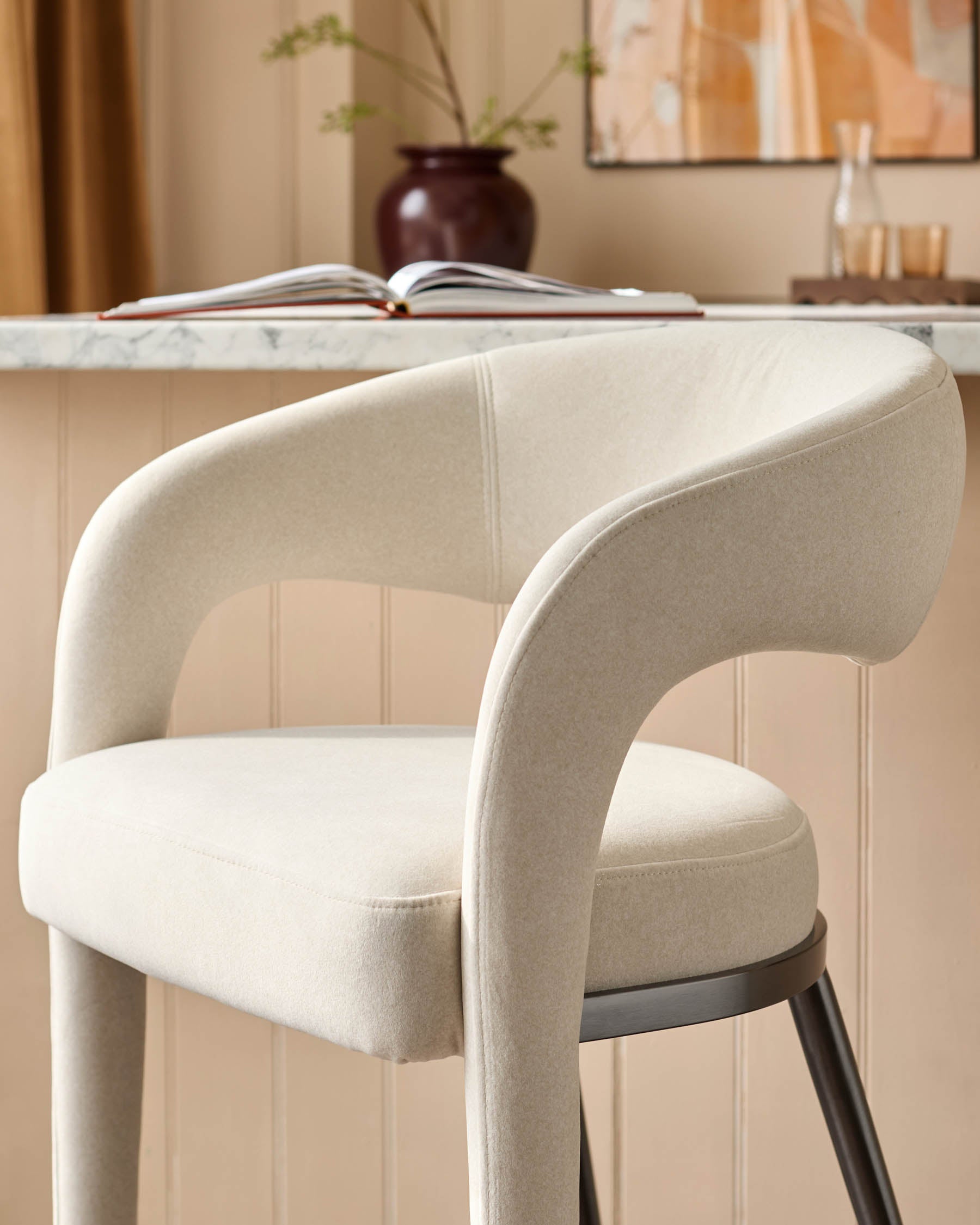 A stylish bar stool with curved arms, upholstered in a light neutral fabric and featuring dark legs.