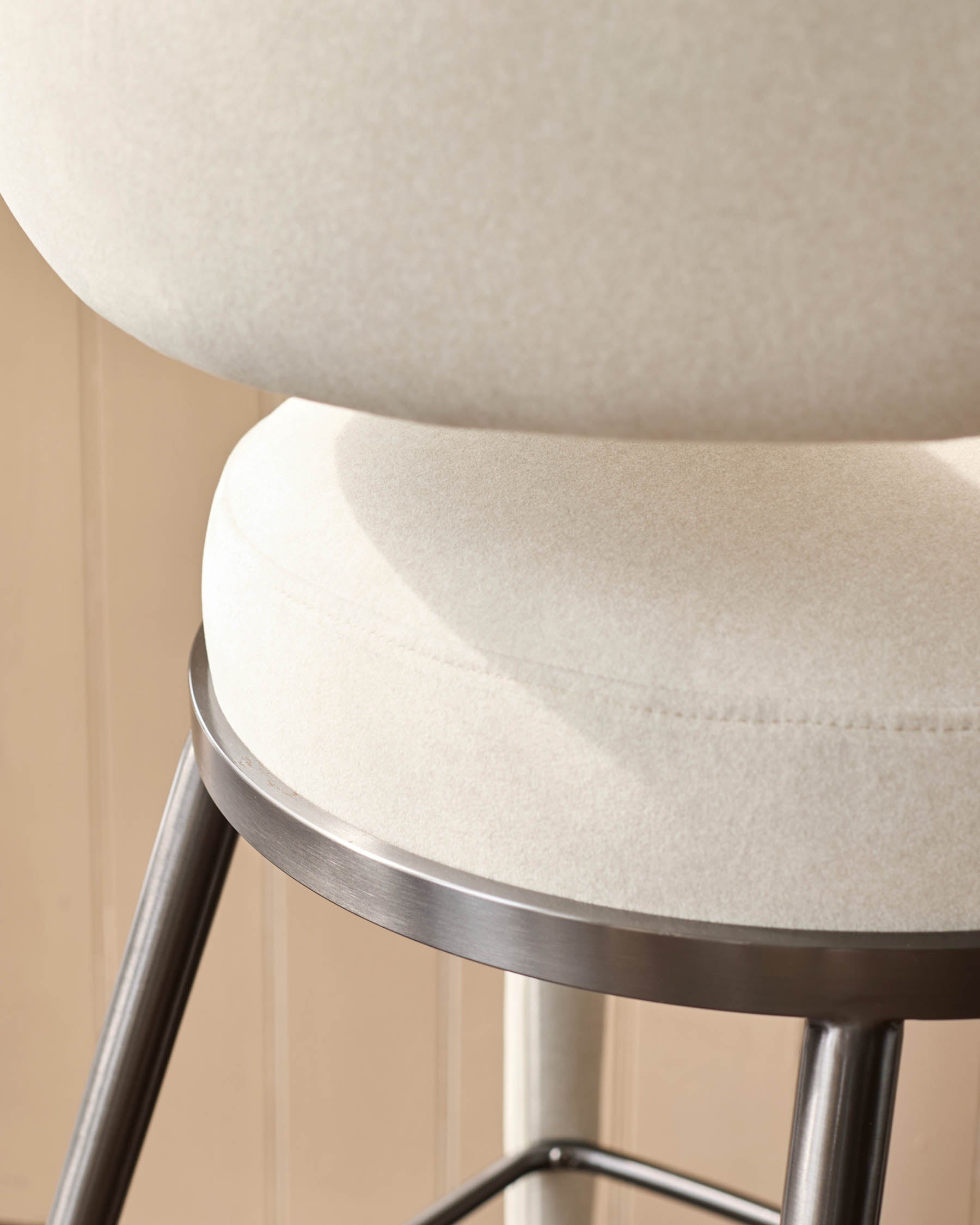 Close-up of a modern stool with a light beige, round cushioned seat and back, supported by a sleek, metallic frame.