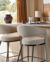 Two neutral-toned bar stools sit at a counter, with a rounded back and dark metal legs.
