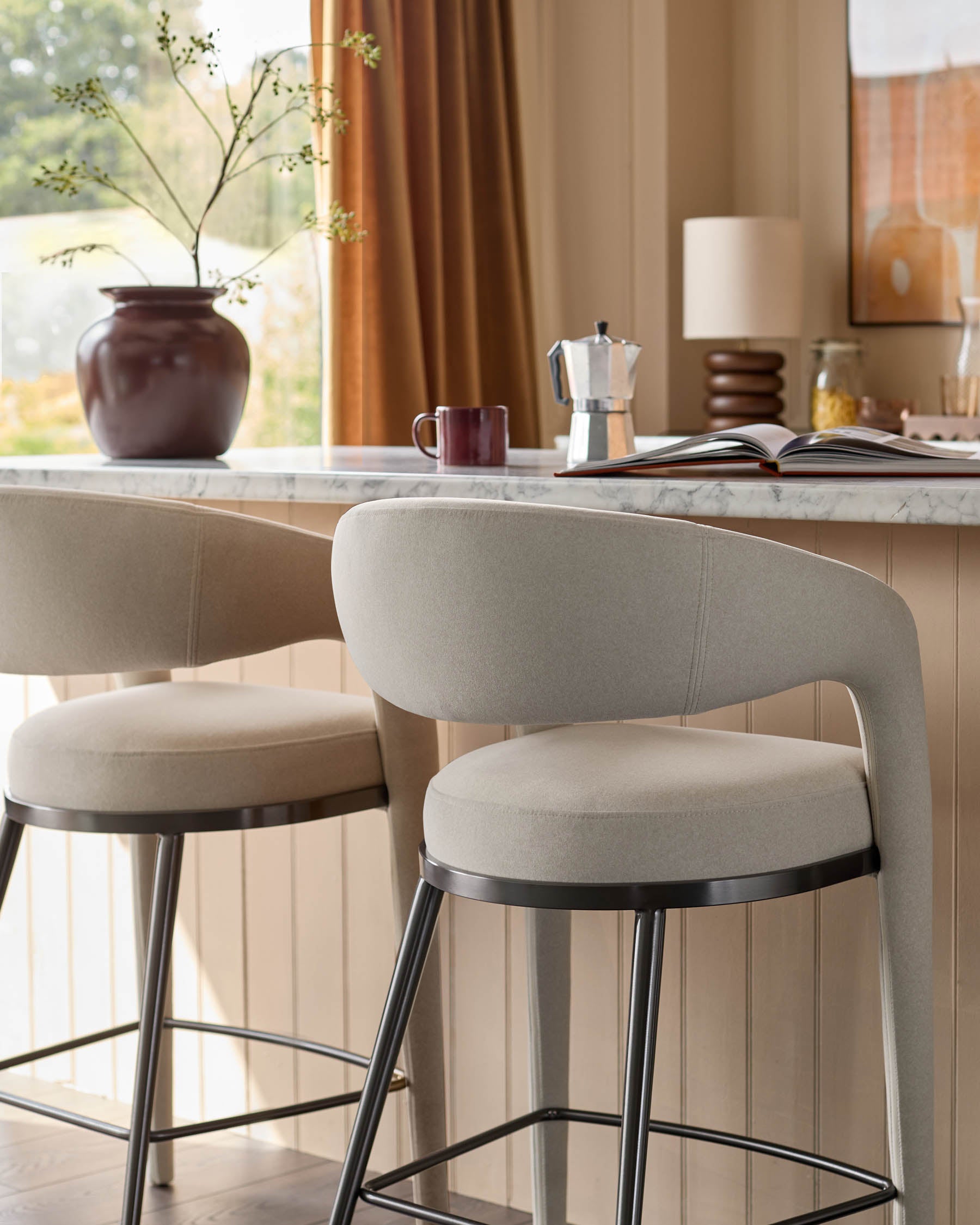 Two neutral-toned bar stools sit at a counter, with a rounded back and dark metal legs.