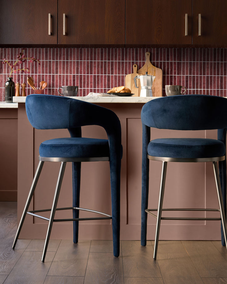 Rhett bar stool in dark navy velvet with curved backrest and brushed metal footrest, shown in kitchen setting.