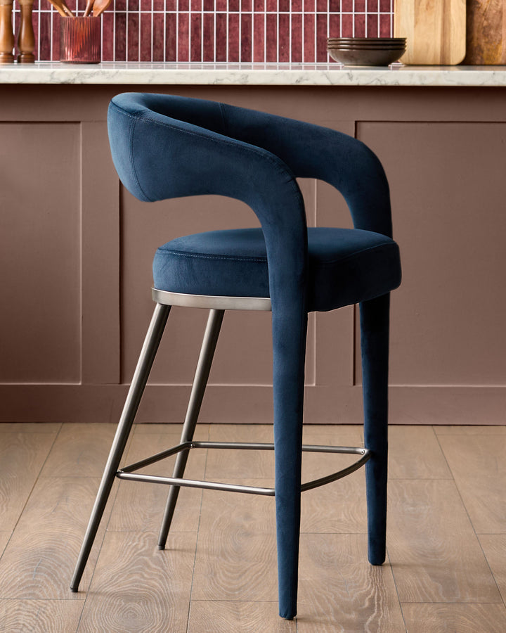 Rhett bar stool in dark navy velvet with curved backrest, metal footrest, and tapered legs in a kitchen setting.