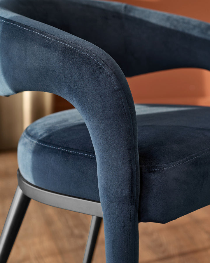 Rhett dining chair in dark navy velvet, close-up of curved armrest and metal frame detail on wooden floor.