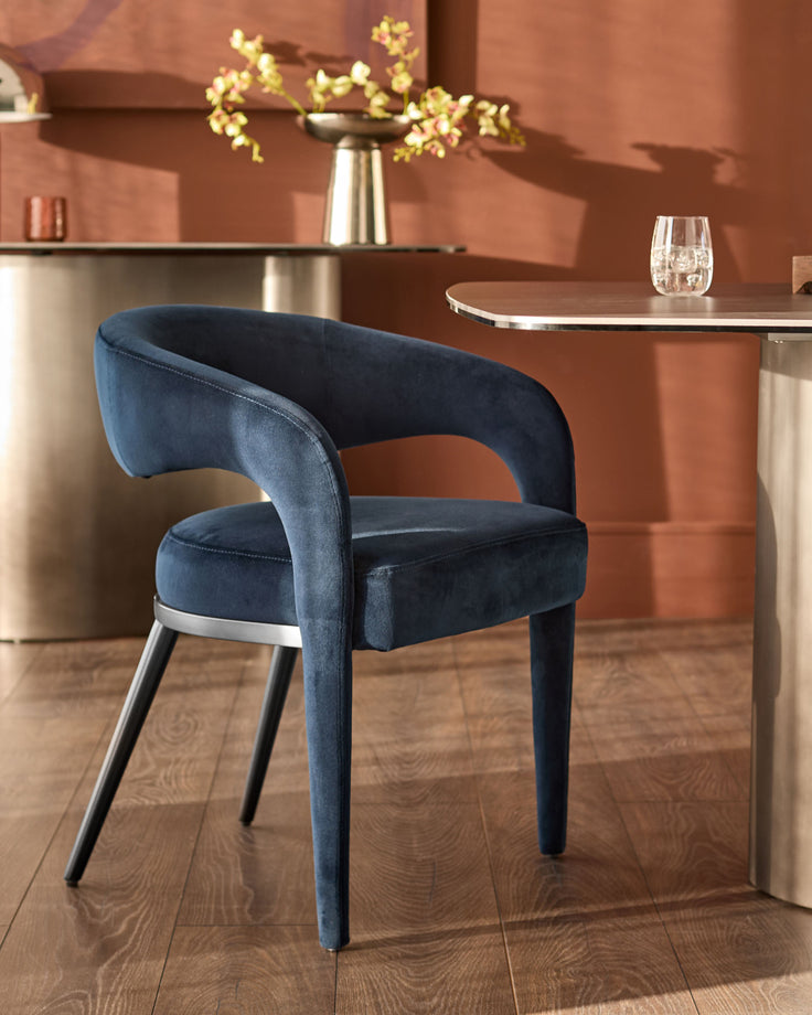 Rhett dining chair in dark navy velvet with curved backrest, metal legs, and chrome accent band, shown in room setting.