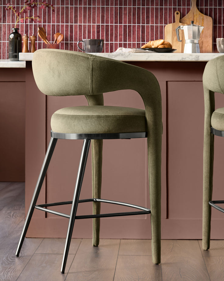 Rhett bar stool in olive green fabric with curved backrest, metal footrest, and tapered legs at a kitchen counter.