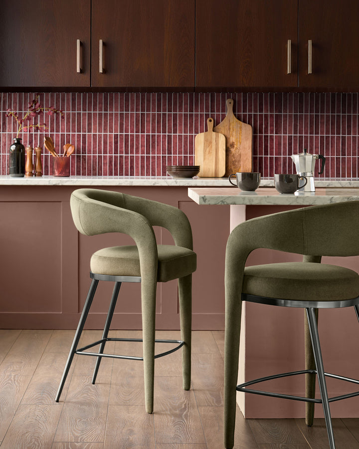 Rhett bar stool in olive green fabric with curved back, black metal footrest and legs, shown at kitchen island.