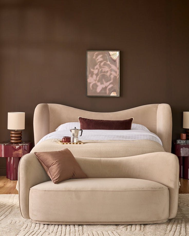 Rhiannon accent sofa in champagne velvet with curved silhouette, displayed in styled bedroom setting.