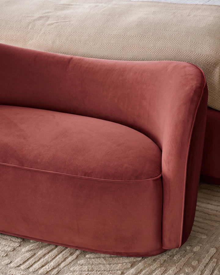 Rhiannon accent sofa in toffee red velvet, side angle view showing curved armrest and seat cushion on patterned rug.