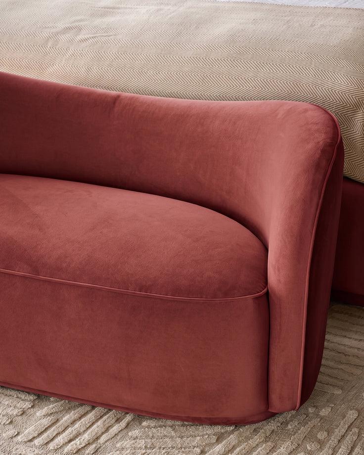 Rhiannon accent sofa in toffee red velvet, side angle view showing curved armrest and seat cushion on patterned rug.