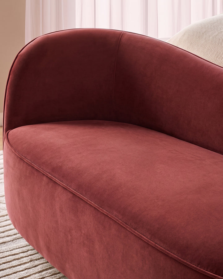 Rhiannon accent sofa in toffee red velvet, curved design with rounded backrest, shown at side angle in modern interior setting.