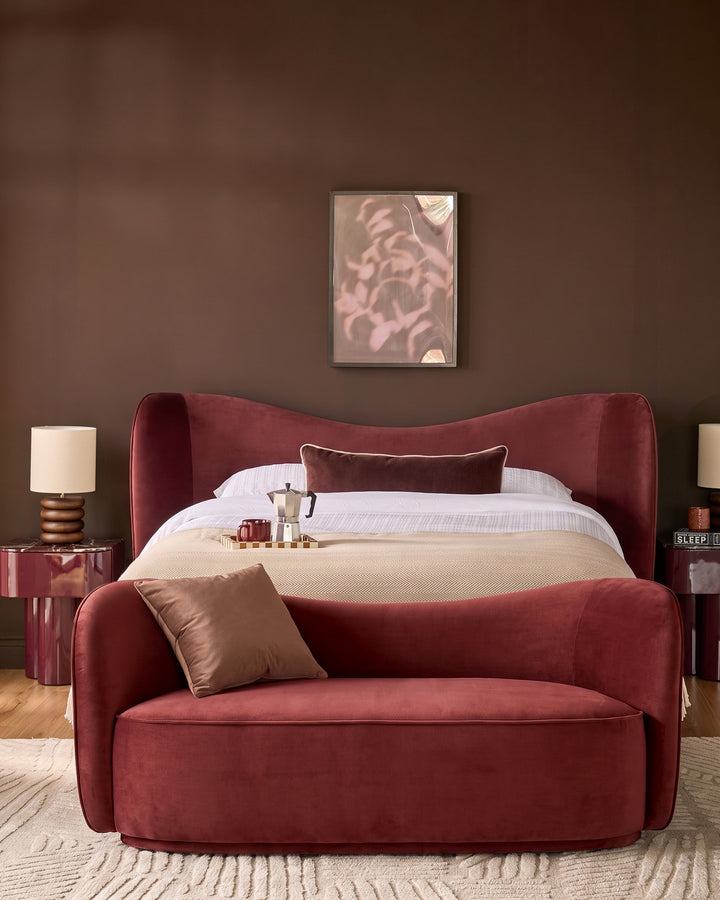 Rhiannon accent sofa in toffee red velvet, curved design, styled in bedroom setting with pillows.