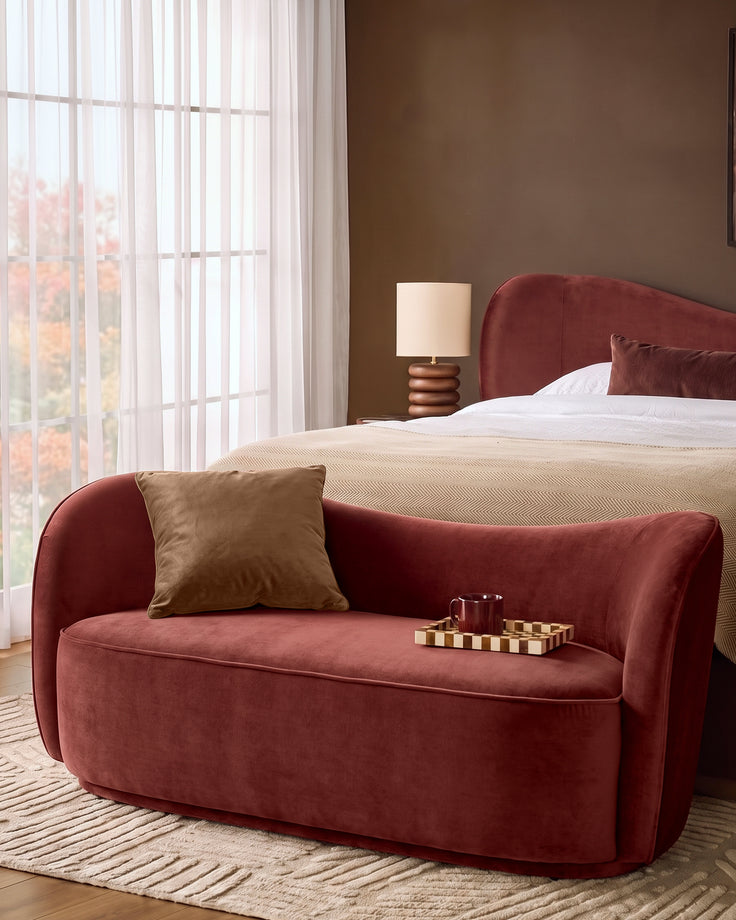 Rhiannon accent sofa in toffee red velvet with curved arms, shown in a bedroom setting with a gold cushion and decorative tray.