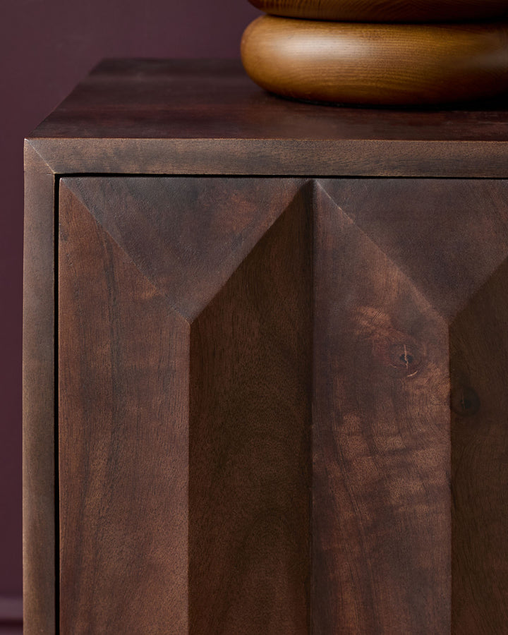 Dark mango wood bedside table with geometric carved drawer front and wooden handle detail, angled view.