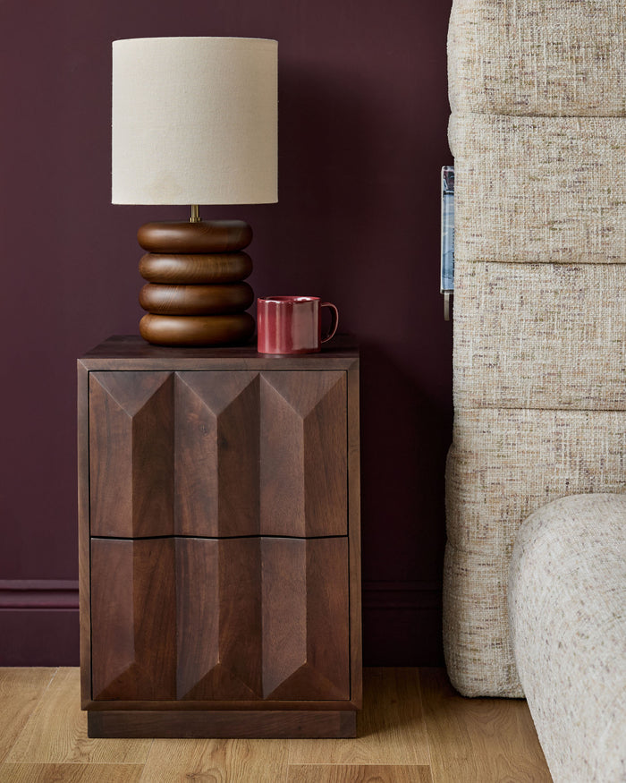 Dark mango wood bedside table with geometric panel doors, displayed with a wooden stacked lamp and burgundy mug.