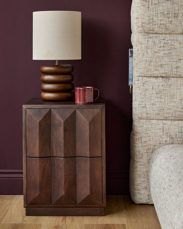 Dark mango wood bedside table with geometric panel doors, displayed with a wooden stacked lamp and burgundy mug.