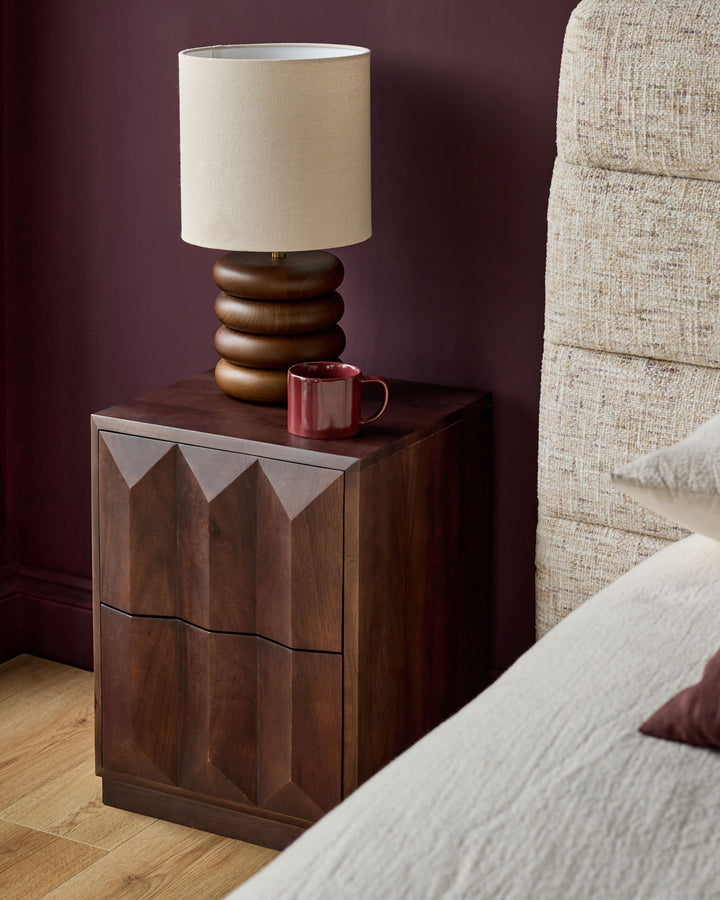 Ria bedside table in dark mango wood with geometric panel doors, styled with a stacked wooden lamp and red mug beside a cream sofa.